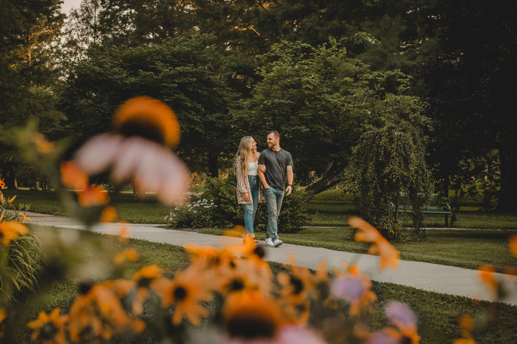 Engaged couple walking along sidewalk in Francis Park STL framed by orange wildflowers.