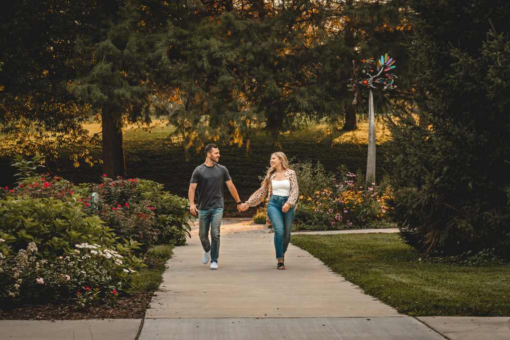 Engaged couple holding hands and laughing while walking down the sidewalk in Francis Park during golden hour.