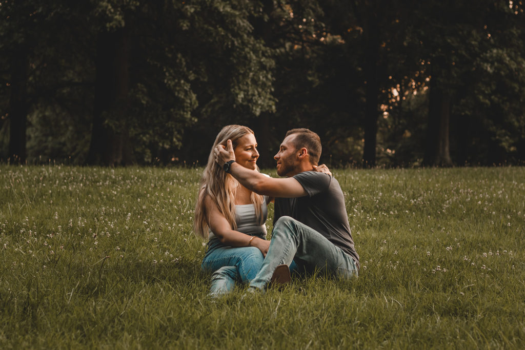 Young man gently pushes his fiance's hair back behind her ear as they sit on the ground.