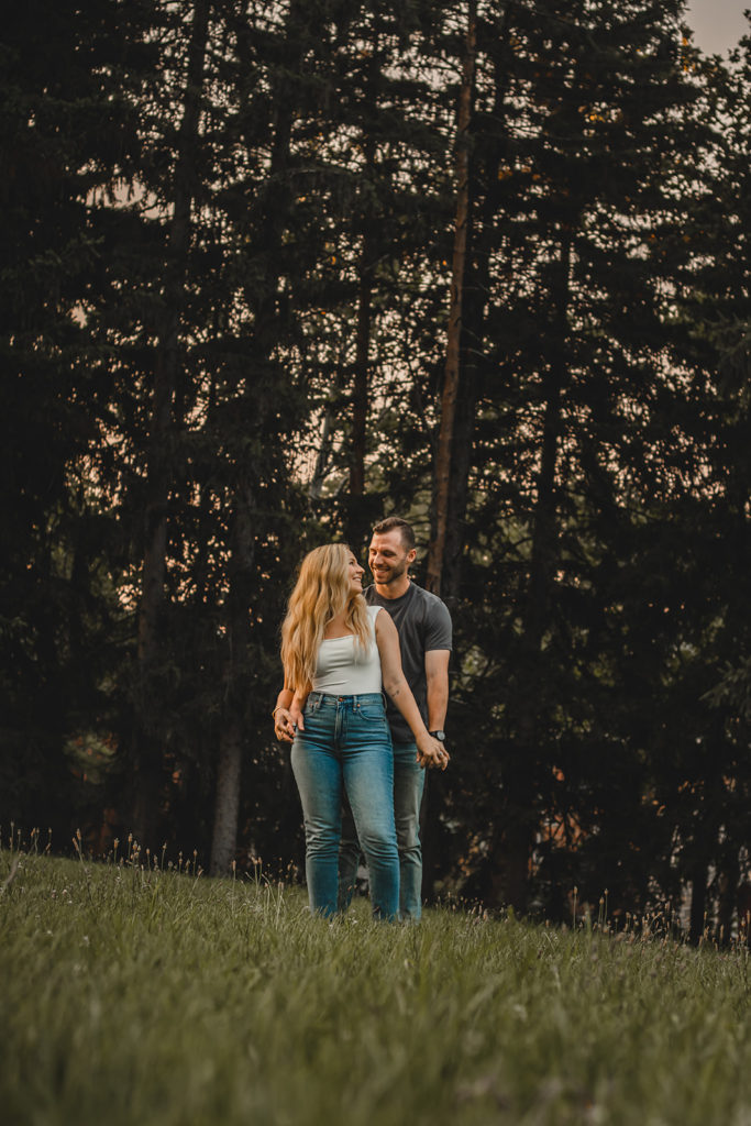 Engaged couple standing in tall grass at a park with trees surrounding them.