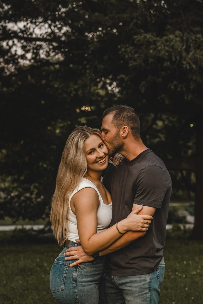 A young woman smiles to camera as her fiance sweetly kisses her cheek.