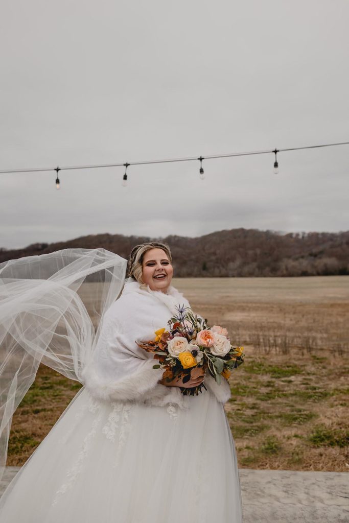 Bride smiling and holding wedding bouquet in front of field under string lights.
