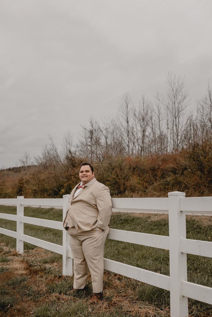 Groom in tan suit standing against white picket fence