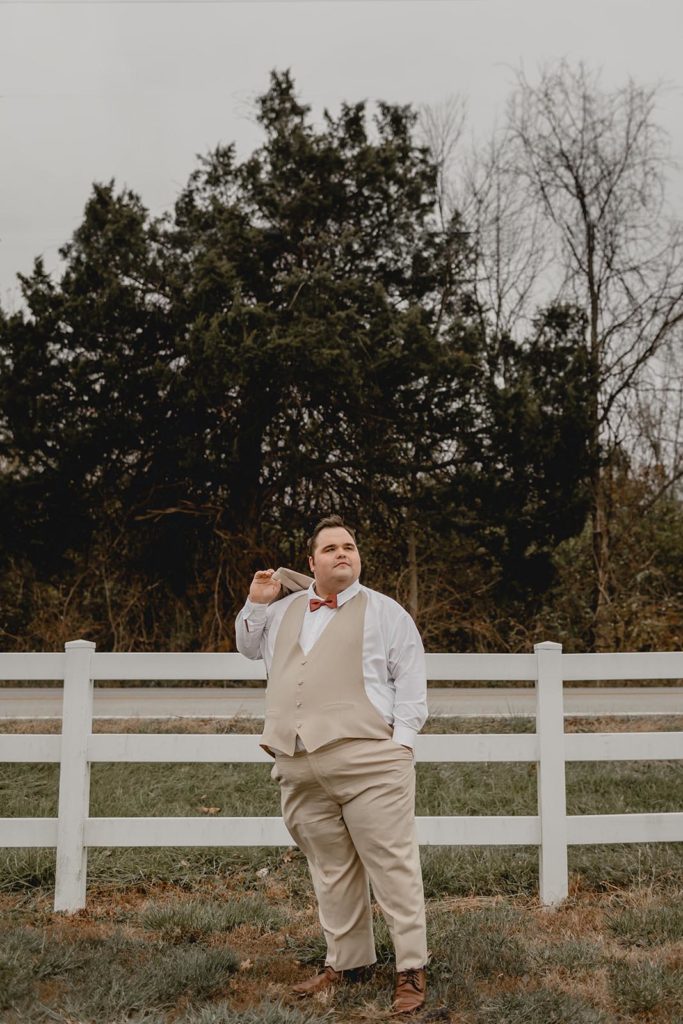 Groom in tan suit standing against white picket fence with jacket over his shoulder