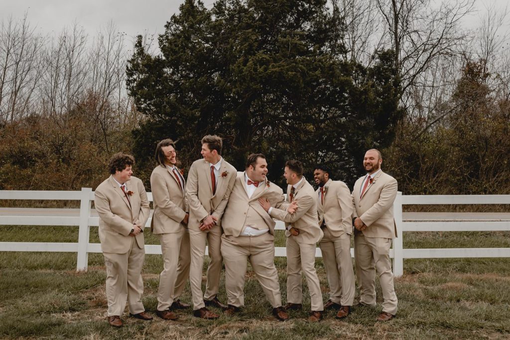 Groomsmen and groom laughing in front of white picket fence in matching tan suits