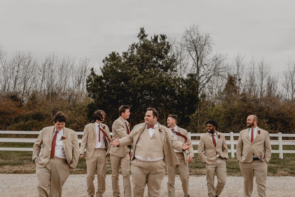 Groom and his groomsmen walking in front of white picket fence in matching tan suites