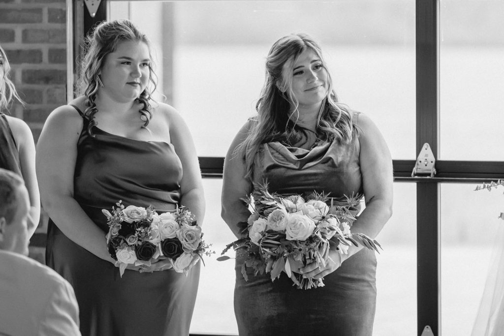 Bridesmaid holding flowers smiling at bride and groom during ceremony