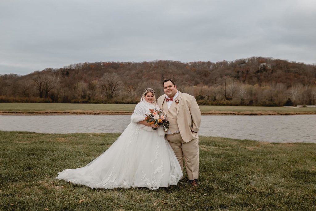 Bride in shaw and groom in tan suite posing in front of a pond