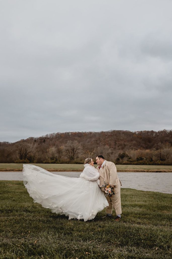 Bride and groom embrace in front of pond while wind blows brides dress