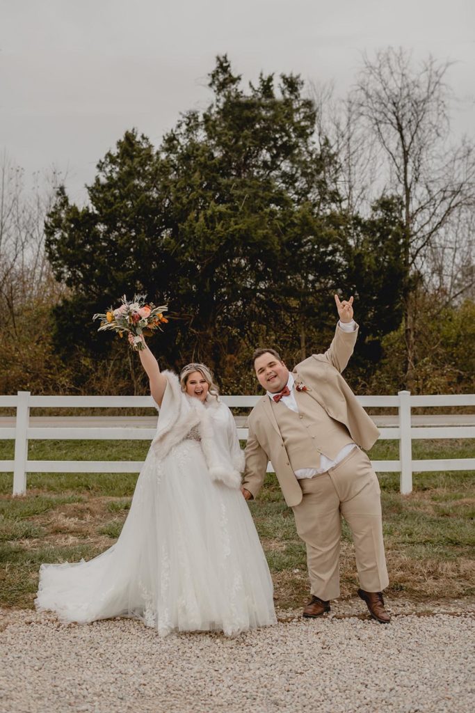 Bride and groom celebrating in front of white picket fence by throwing their hands in the air