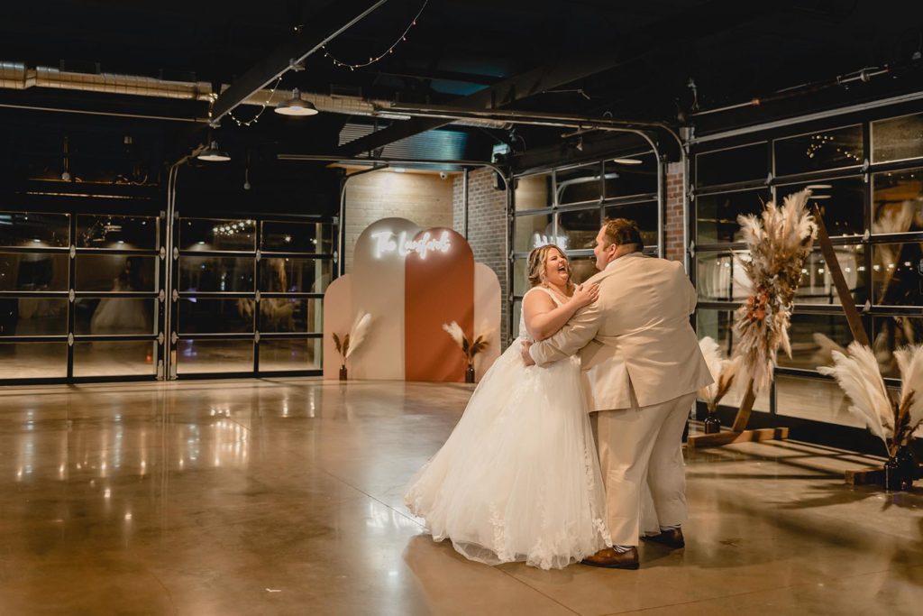 Bride and groom dancing to their first song after getting married on sealed concrete floor with neon sign background.