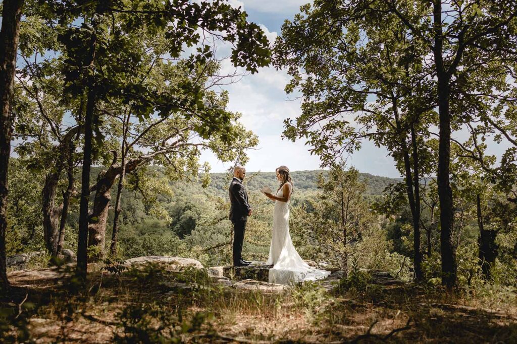 Wide centered shot of bride reading from her vow book to her husband between a wooded outcropping