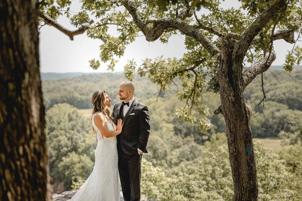 Bride and groom lovingly gaze towards each other, perfectly centered between two trees above the forest top on Thinking Rock at Lost Hill Lake
