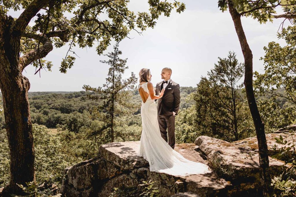 Wide shot that features the back of a bride's dress as she faces her groom on top of a large rock perched at the edge of Lost Hill Lake's Thinking Rock scenic overlook