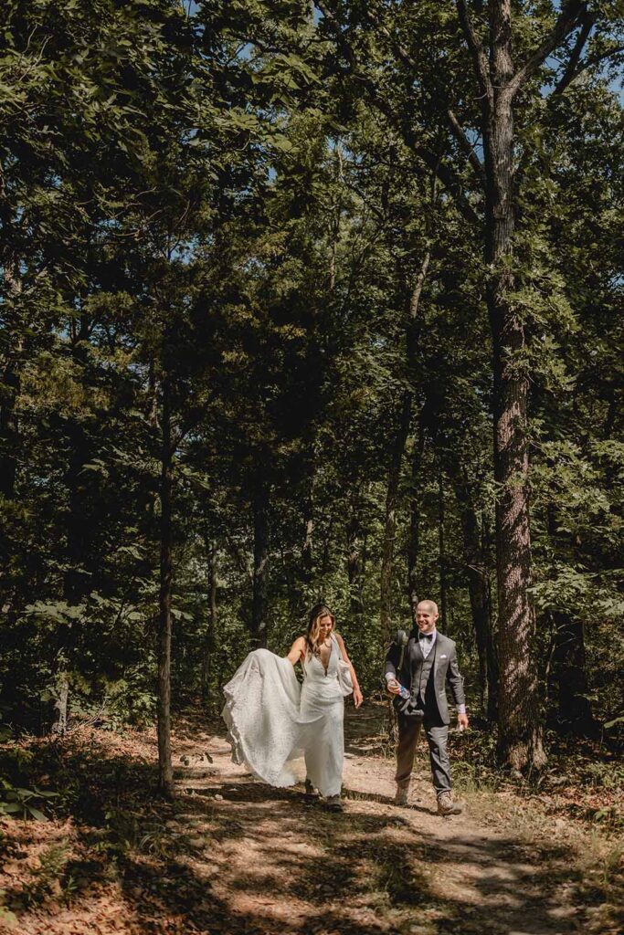 Wide shot of bride and groom laughing together as they haul their gear and hike along a forest path