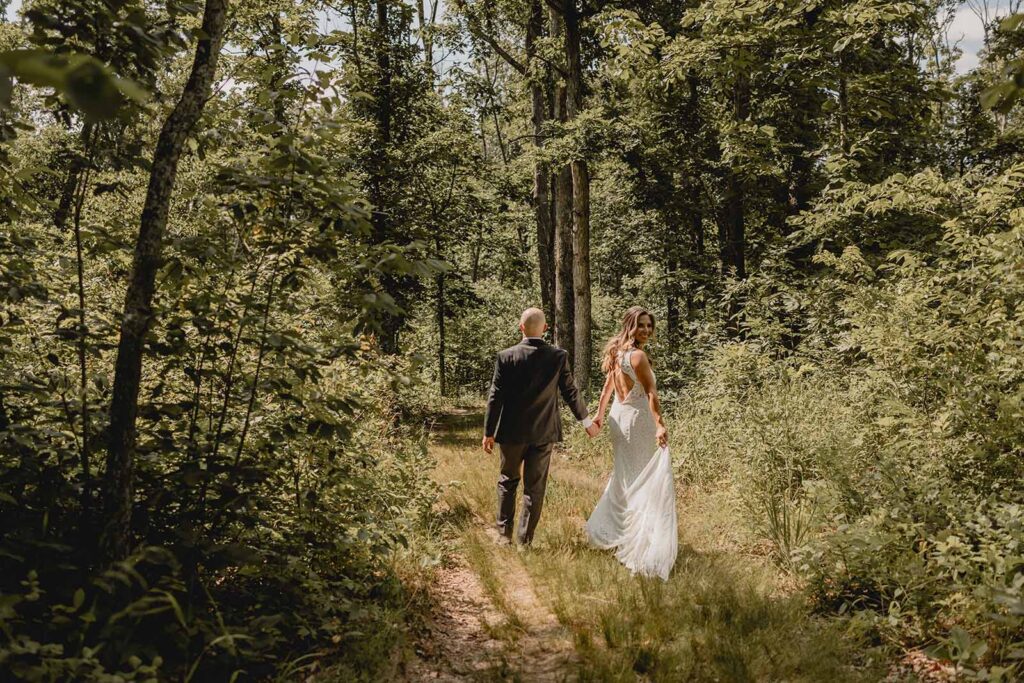 A groom guides his wife by the hand as she looks back towards the camera over her should in a green forest clearing