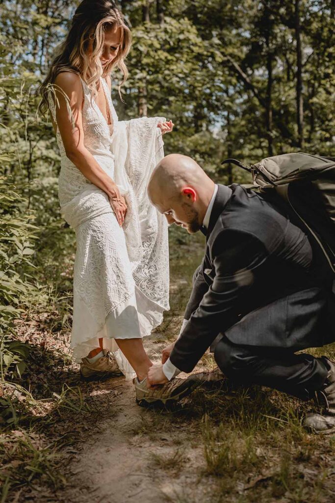 Close up of a bride pulling up her lace wedding dress for her groom in a black suit to kneel and tie her hiking shoes