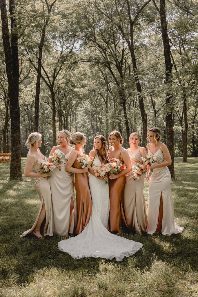 Full body shot of centered bride surrounded by her 6 bridesmaids candidly laughing with their bouquets