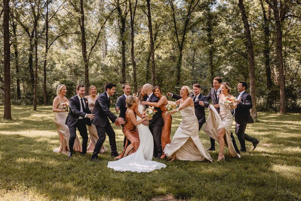 Bride and groom kissing as their wedding party rushes in from either side for a giant group hug in a walnut grove