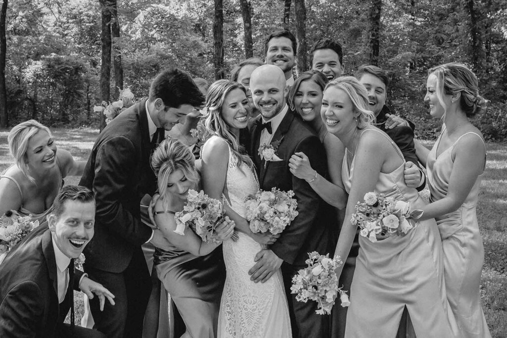 Black and white close up of bride and groom smiling as their wedding party surrounds them for a group hug
