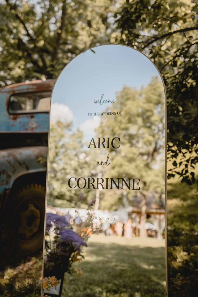 Close up of a rounded top oval mirror sitting outside with text that reads "Welcome to the wedding of Aric and Corrinne"