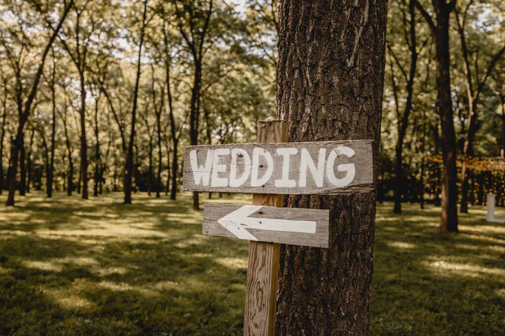 A wooden painted sign that reads "Wedding" with an arrow leans against the trunk of a tree
