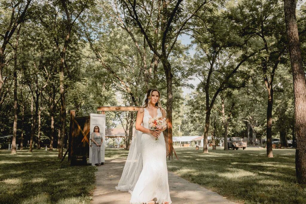 Bride walks wildflower bouquet down stone aisle in a green-covered walnut grove with speckled sunlight