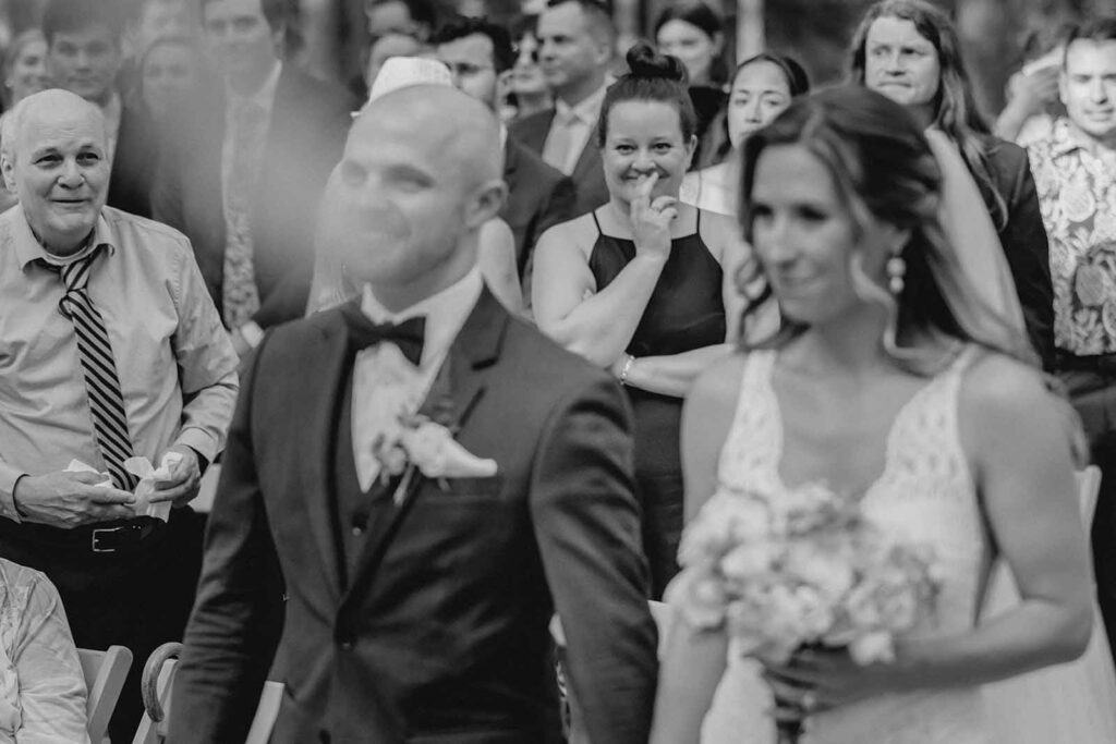 Black and white shot looking past out of focus bride an groom at delighted wedding guests watching them walk down the aisle