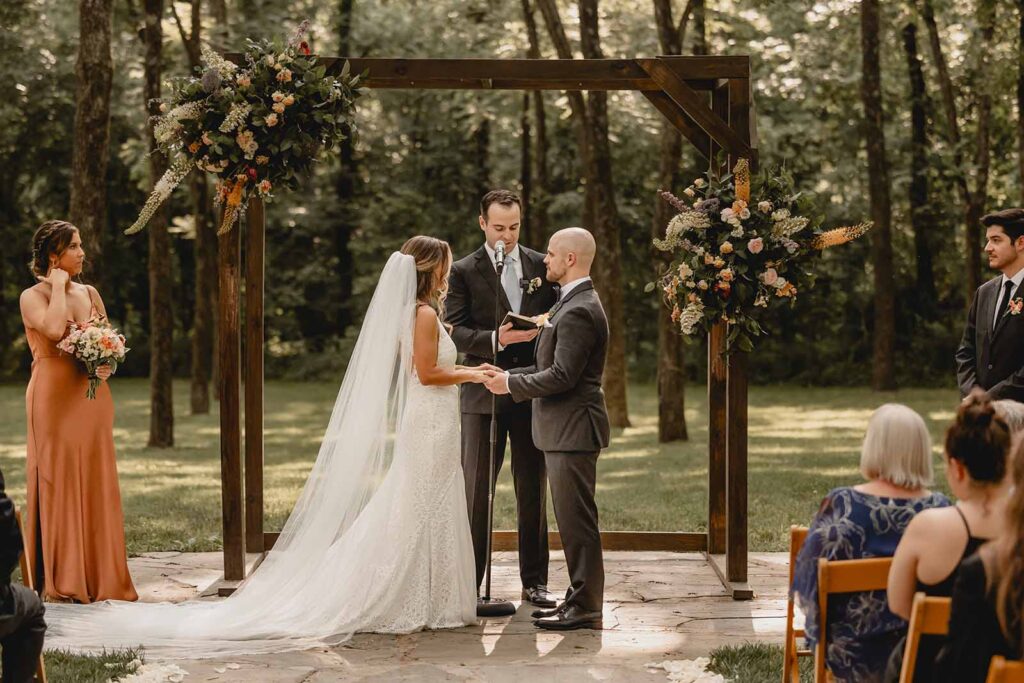 Bride and groom holding hands under a wooden rectangle arbor as officiant begins their outdoor summer ceremony