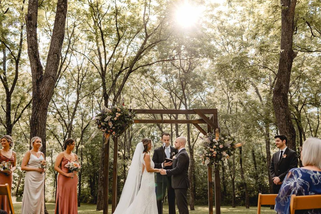 A sunflare shines through a canopy of green tall trees over a bride and groom in front a wooden arbor with officiant and wedding party
