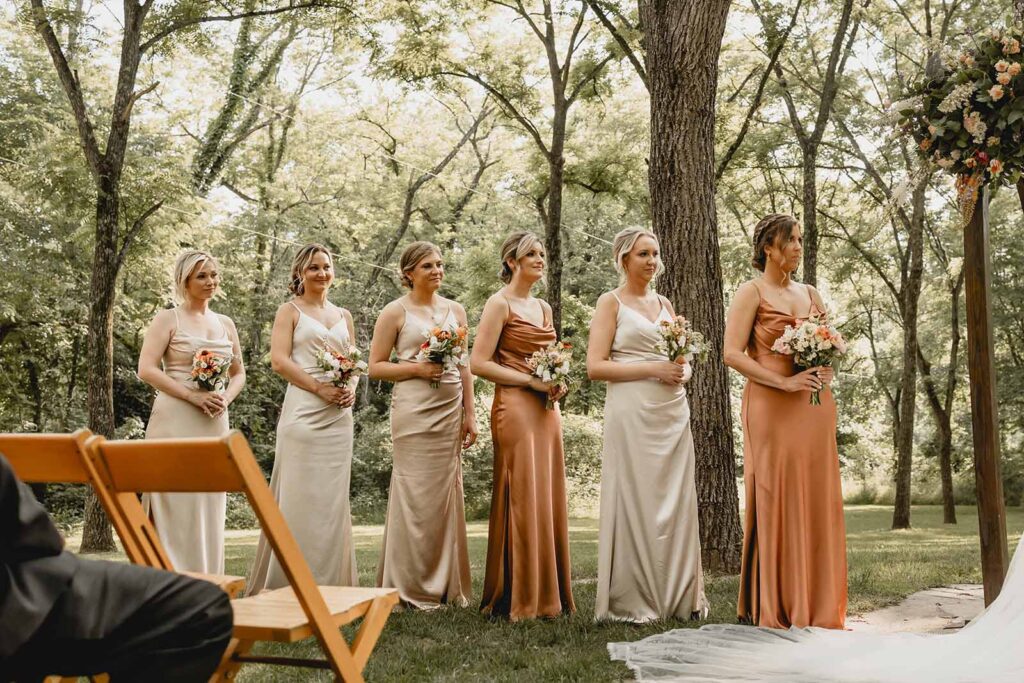 Bridesmaids in a mixing of neutral creams and burnt orange hold bouquets and look on towards a wedding ceremony