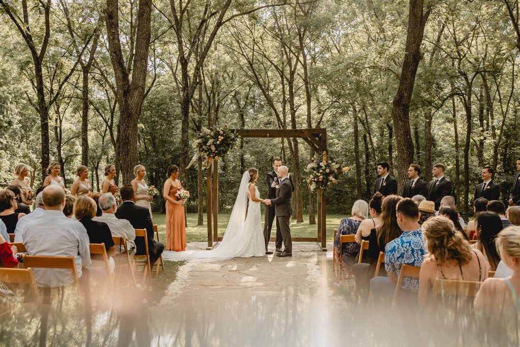 Mid-crop shot of centered bride and groom holding hands under their arbor flanked by their wedding party in the walnut grove ceremony