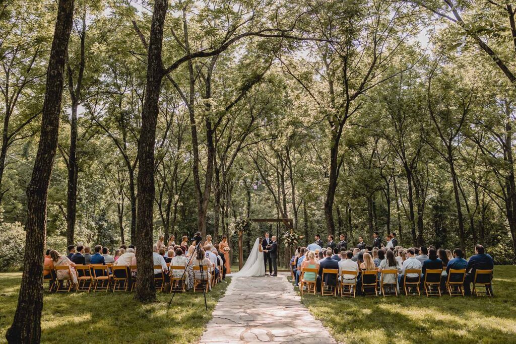 Wide centered shot of full ceremony setup beneath a grove of tall trees and summer foliage