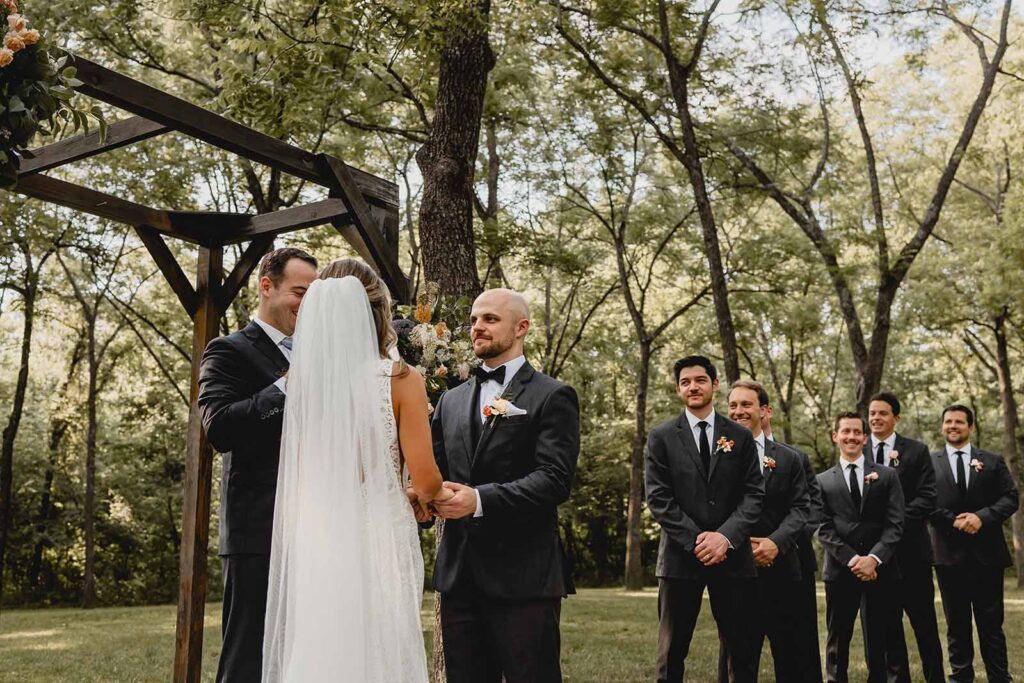 Groom smiles holding his bride's hands as a line of groomsmen smile behind, shot from the perspective behind the bride