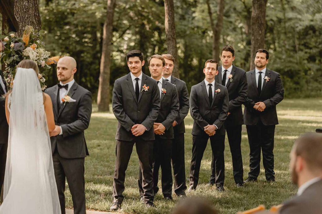 Line of groomsmen in black suits with wildflower boutonniere looking on at their friend's ceremony
