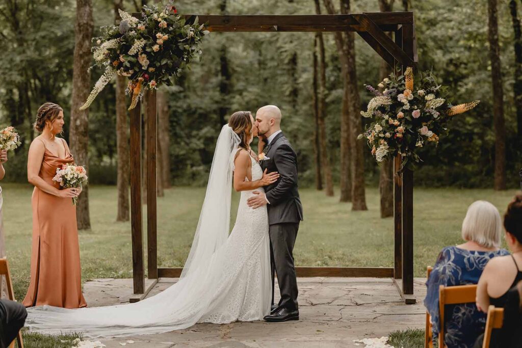 Close up shot of a bride and groom kissing in a green grove under a wooden square arbor