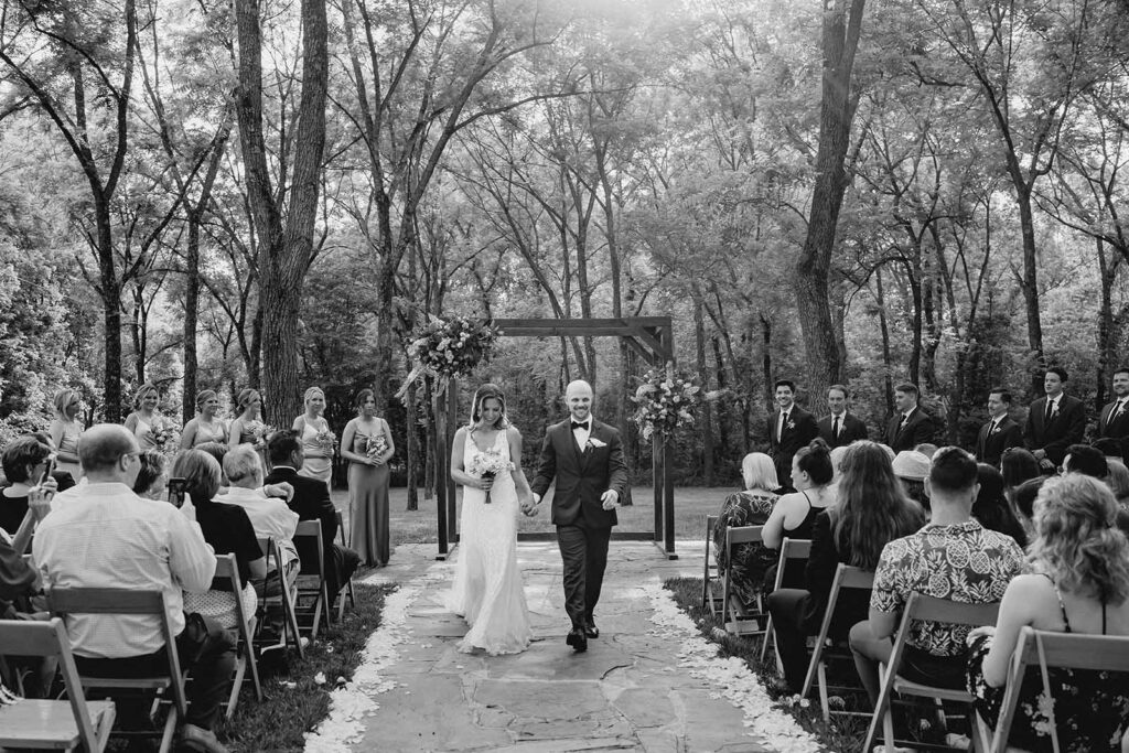 Black and white shot of boho bride and groom walking down the stone aisle lined with white rose petals in an outdoor ceremony