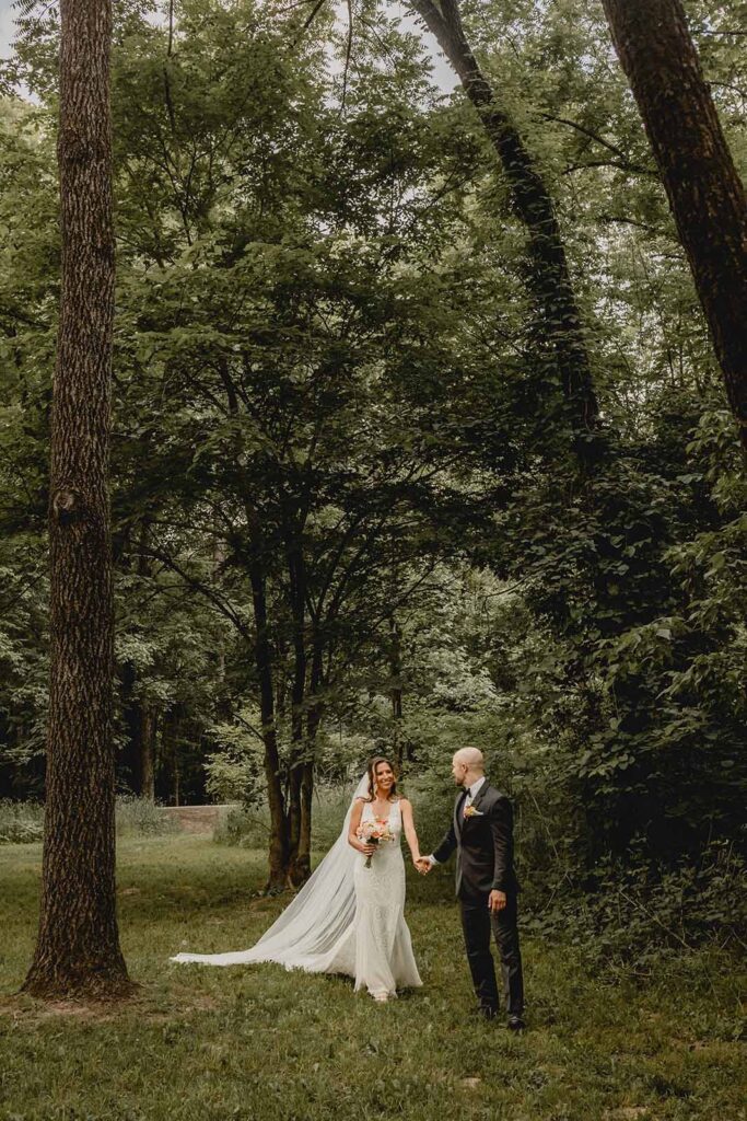 Far shot of bride in long veil holding wildflower bouquet and husbands hand as he guides her through a green tree covered meadow