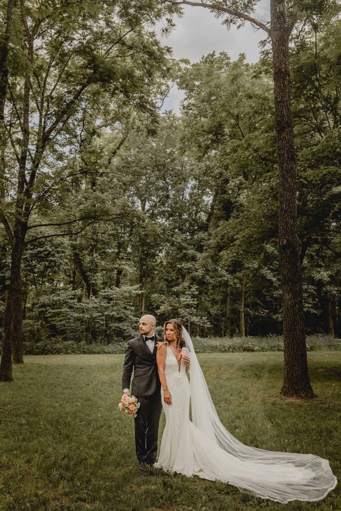 Full body far shot of bride and groom centered between two tall trees looking out in an editorial pose with dramatic long cathedral veil 