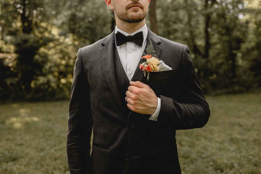 Groom detail closeup of his ring hand holding onto his label, black suit and bowtie and colorful wildflower boutonniere