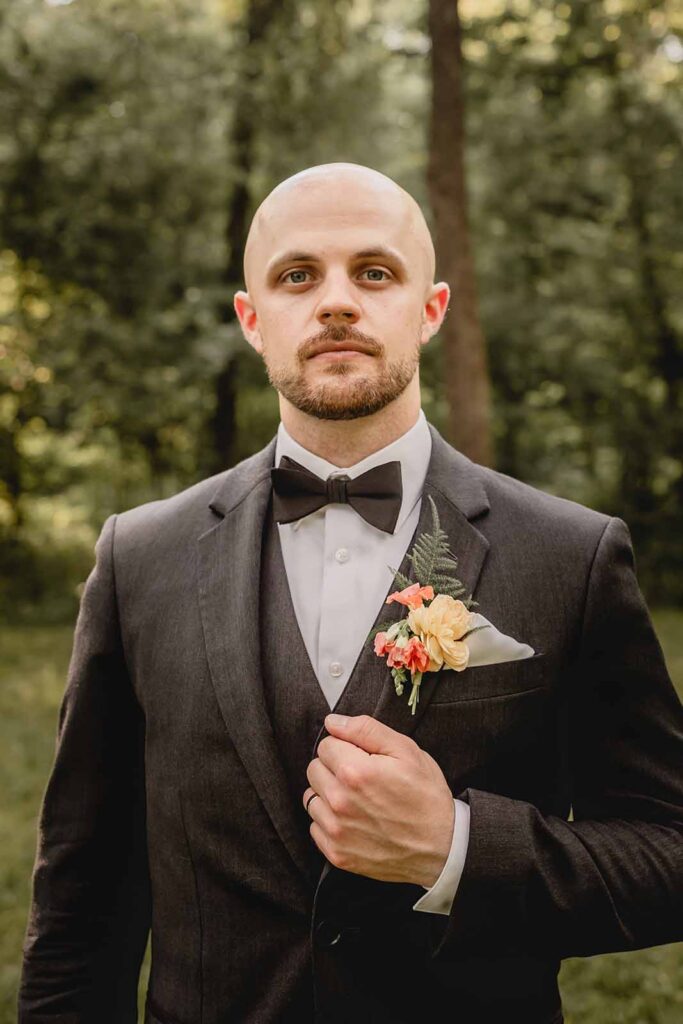 Headshot of young bald groom in black suit and bowtie with wildflower boutonniere