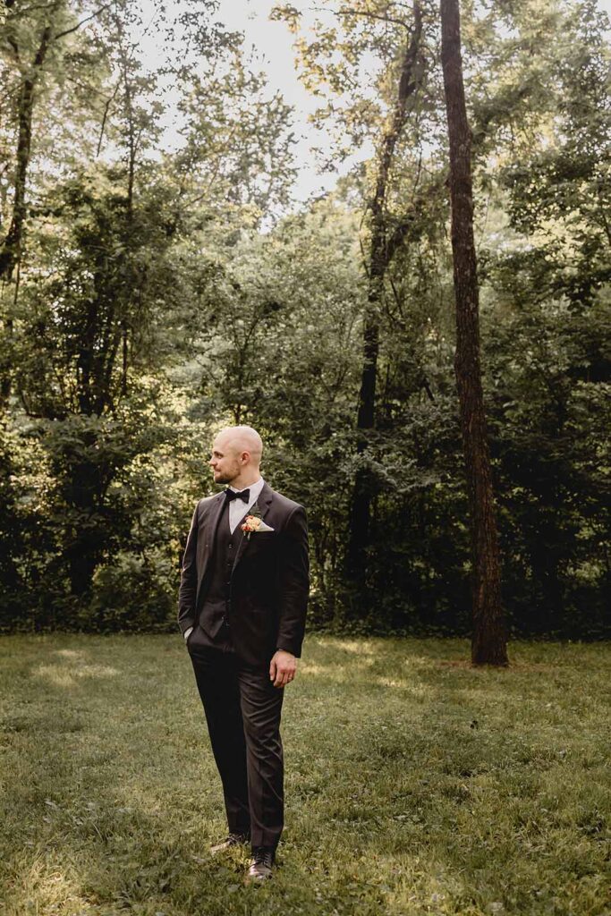 Full body shot of young bald groom in black suit and bowtie with wildflower boutonniere in secluded grove of greenery