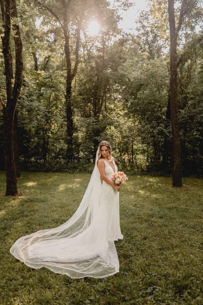 Full body shot of young brunette bride in lace wedding dress with a long train and veil with wildflower bouquet in secluded grove of greenery