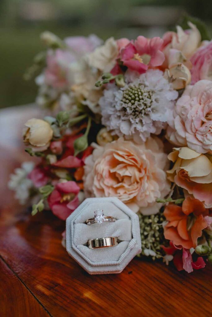 Gold wedding rings in a cream ring box lean against a bouquet of peach, white, pink, and orange wildflowers
