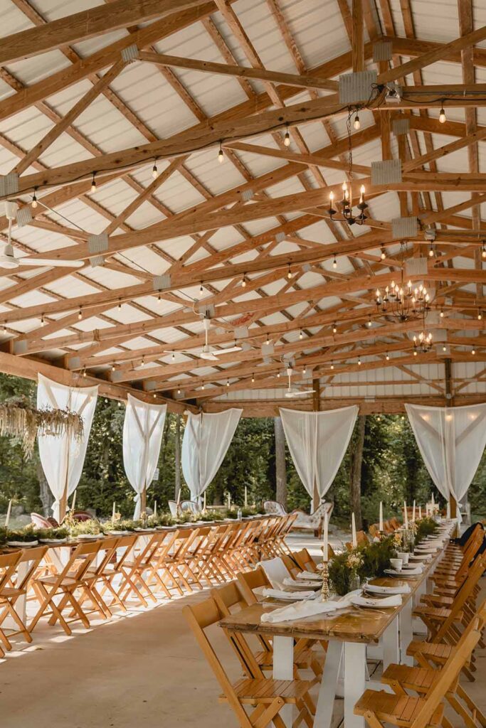 Wide shot of family-style long wedding dinner tables lined with greenery and wooden folding chairs in an wood-frame open pavilion