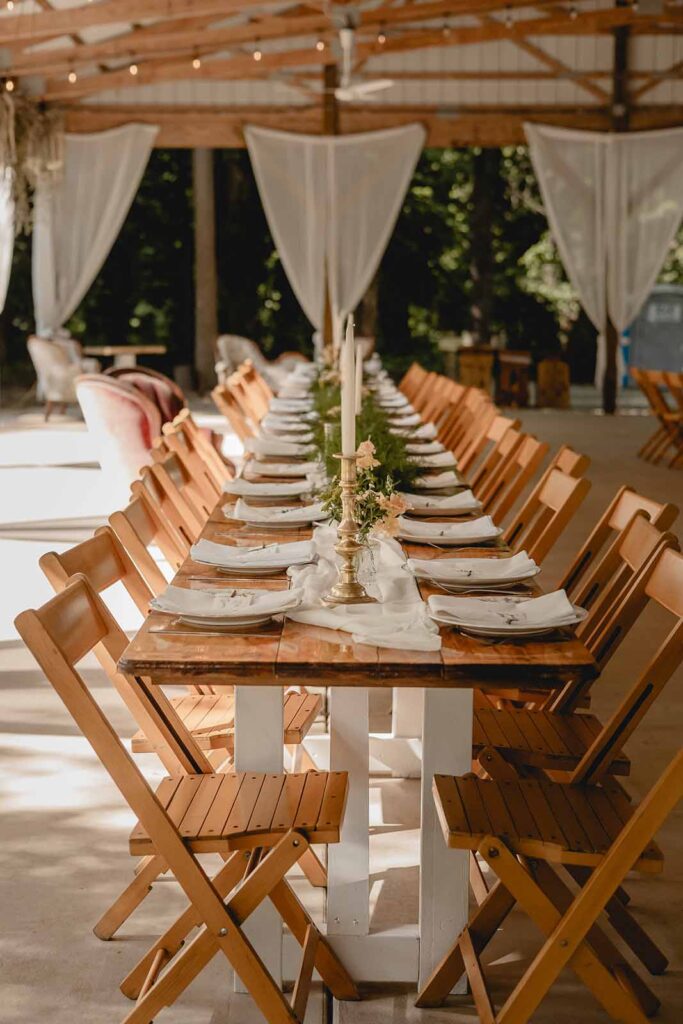 End of a long family-style wooden wedding dinner table with golden candlesticks, long taper candles and strand of greenery down the center
