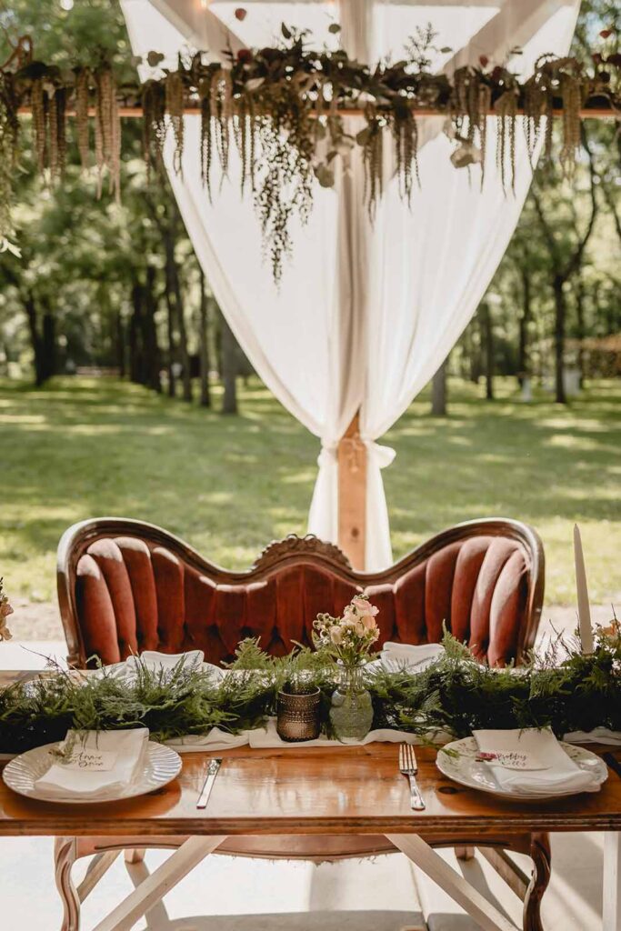 Antique red couch sits beneath a hanging floral arrangement at the head table of a outdoor pavilion wedding reception