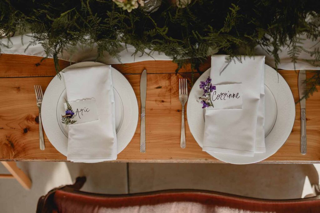 Aerial shot of bride and grooms dinner place settings or white plates and napkins with hand lettered placecards with pressed dried purple wildflowers set on wood table