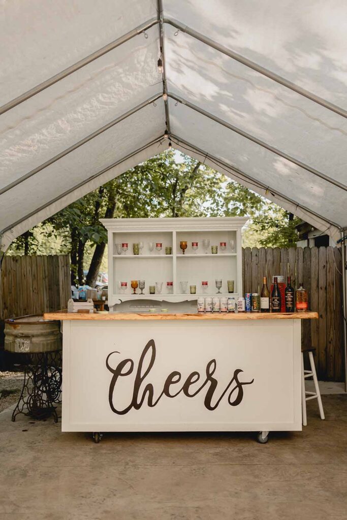 Outdoor shabby chic bar with "cheers" painted in black on the front in a script text and white bookcase of eclectic colored glassware behind