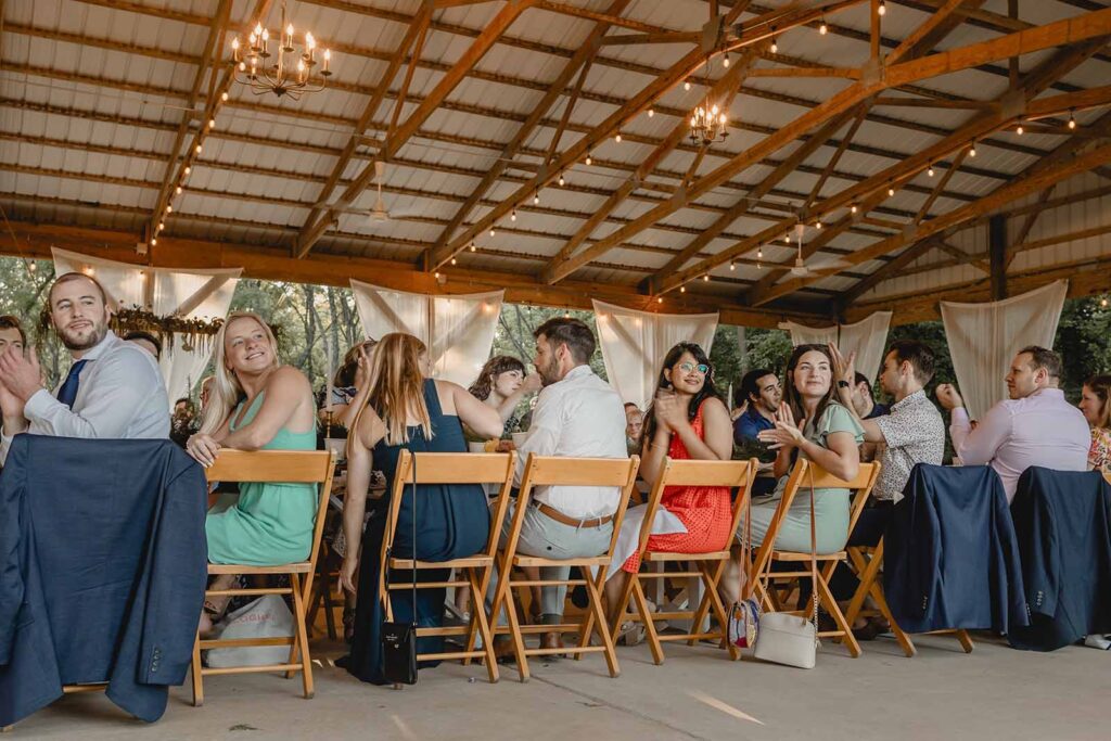 Guests sitting at a long table under wooden pavilion look back over their chair and clap for grand reception entrance